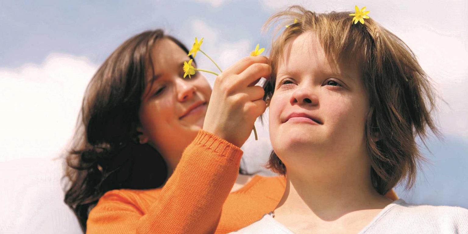 Eine junge Frau steckt einem Mädchen mit Down-Syndrom Blumen ins Haar.