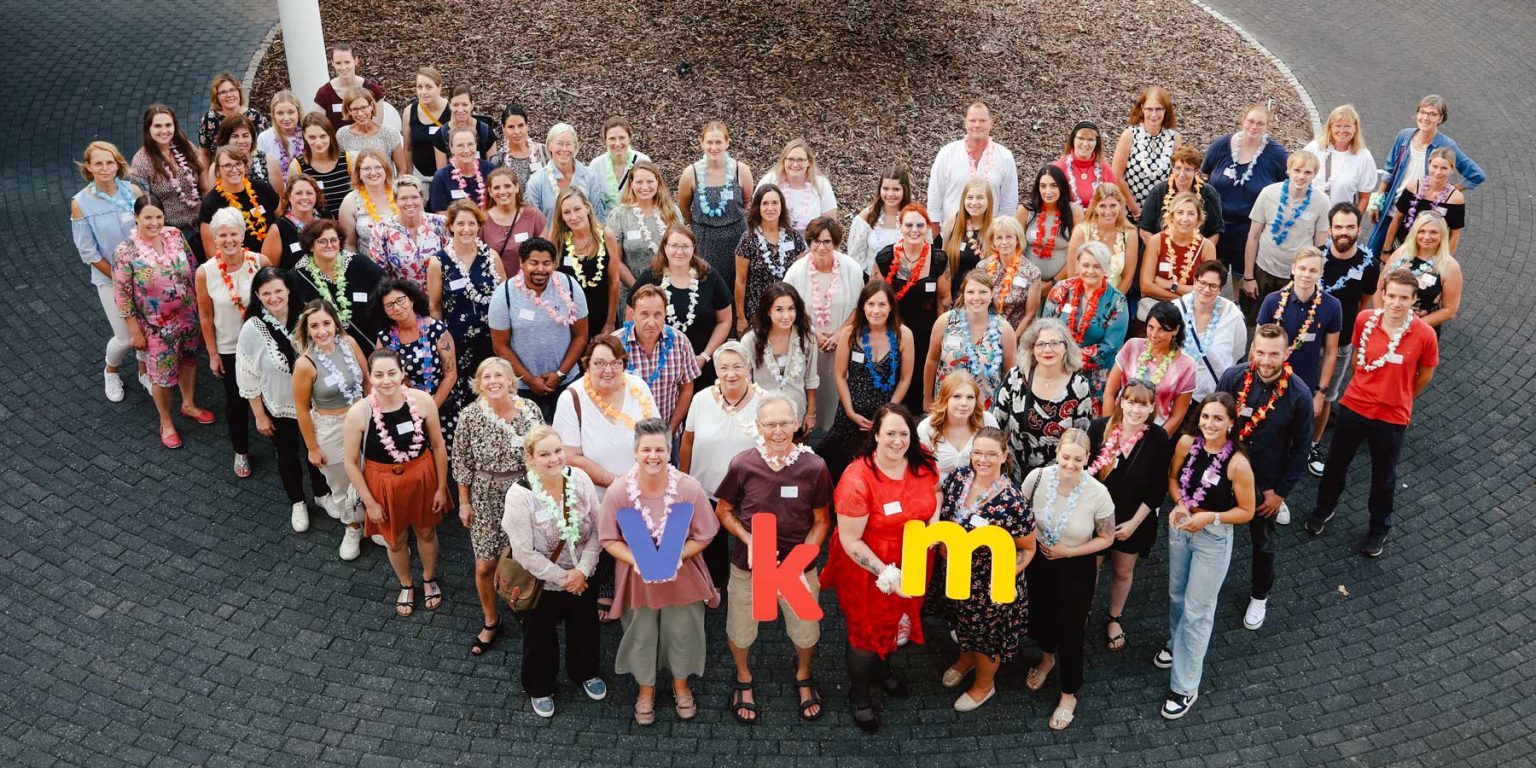 Zahlreiche VKM Mitarbeiter posieren für eine Gruppenaufnahme während eines Sommerfests. Vier Mitarbeitende halten die VKM Buchstaben aus Acrylglas.