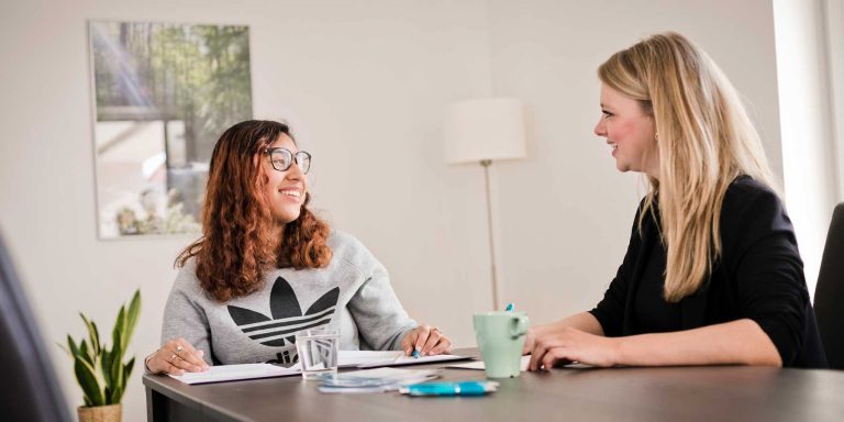 Zwei Junge Frauen sitzen während eines Beratunstermins zusammen am Tisch und lächeln.