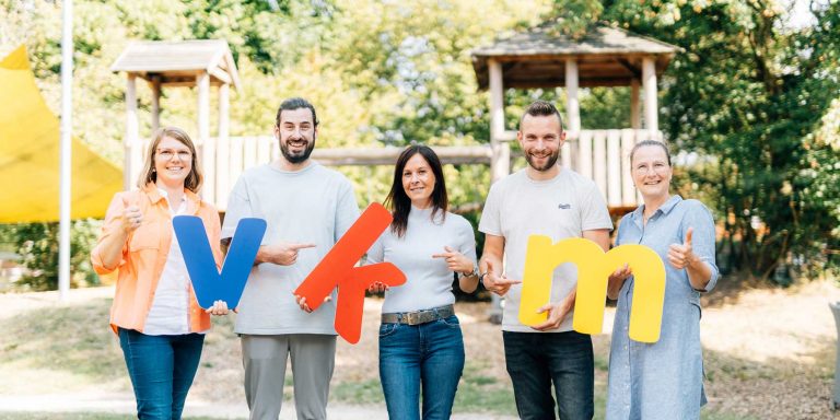 VKM Mitarbeiter posieren für ein Gruppenfoto vor einem Klettergerüst und halten die VKM Buchstaben aus Acrylglas.
