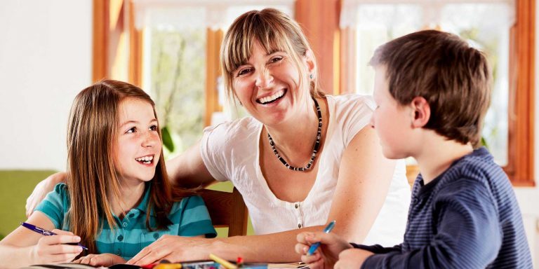 Eine Frau sitzt mit zwei Kindern an einem Tisch. Die Kindern sitzen an ihren Hausaufgaben.