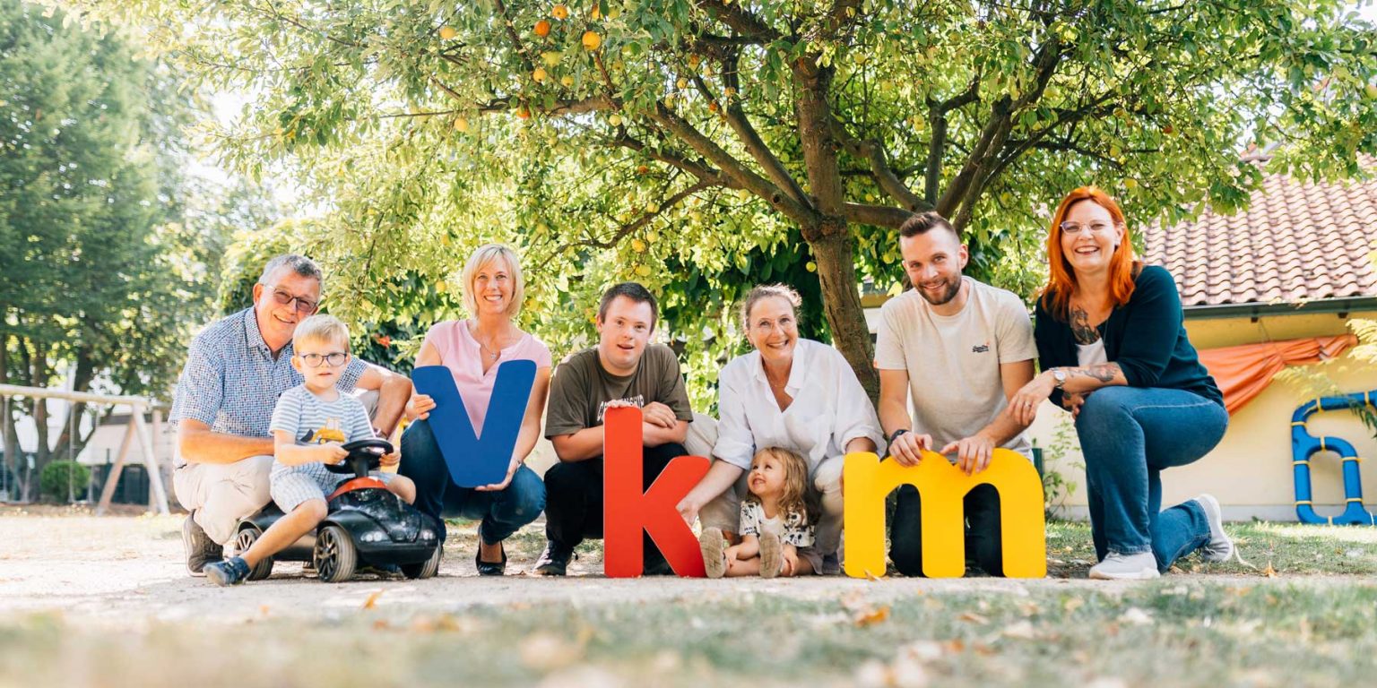 VKM Mitarbeiter, Klienten und Kitakinder posieren vor einem Baum und halten VKM Buchstaben aus Acrylglas.