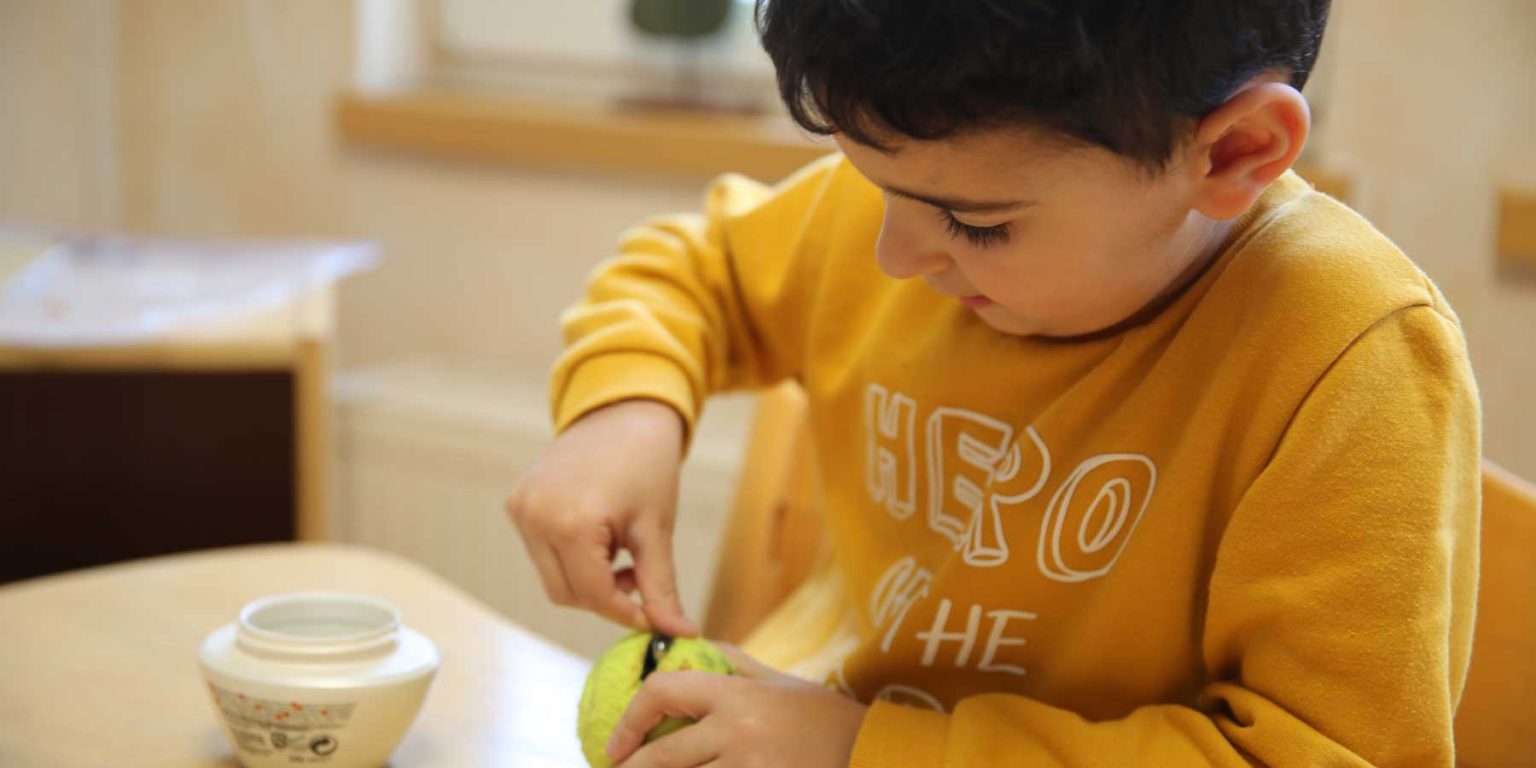 Ein Junge in einem gelben Pullover steckt einen kleinen Gegenstand in einen aufgeschnittenen Tennisball.